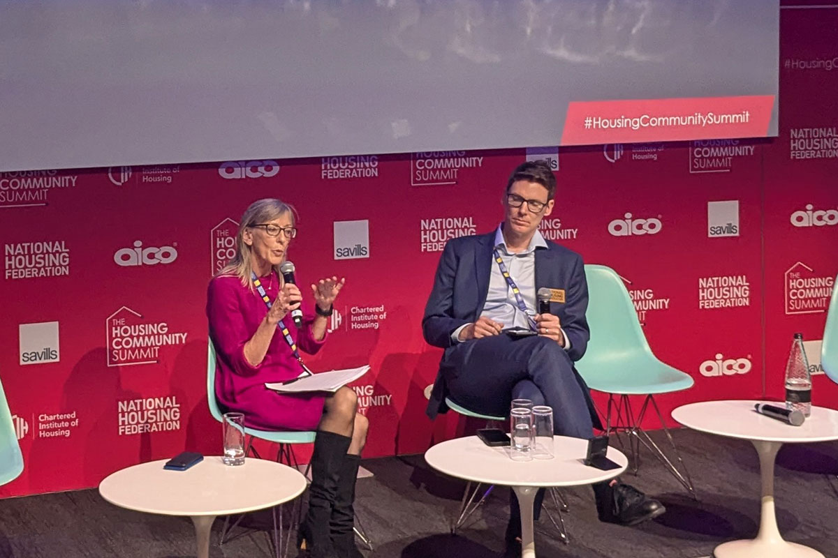 Fiona MacGregor holding a microphone, speaking at an event, sitting next to a man in a suit Fiona MacGregor holding a microphone, speaking at an event, sitting next to a man in a suit
